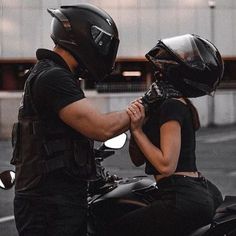 a man and woman on a motorcycle talking to each other while wearing helmets, standing in front of a building