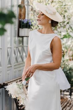 a woman in a white dress and hat standing on a bench holding a flower bouquet