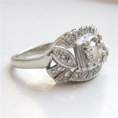 This vintage 1940s ring has a central diamond, weighing approximately 0.50ct (H color, SI2 clarity). An additional 0.25ct of diamonds surrounds the center, and the top of the ring - with the way the mounting is carved and the stones are set - has the illusion of motion. The mounting is made entirely of 14K white gold.SIZE 6.5 (can be sized) Vintage Diamond White Platinum Halo Ring, Vintage Platinum Halo Ring In Diamond White, White Diamond Art Deco Cluster Ring, Vintage Halo Ring With Diamond Accents, Vintage White Halo Ring With Prong Setting, Vintage Platinum Halo Ring With Brilliant Cut, Classic Cluster Ring With Halo Setting, Vintage Diamond White Halo Ring, Vintage White Diamond Ring With Halo Setting