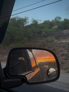 the rear view mirror of a car as it drives down a road with trees and hills in the background