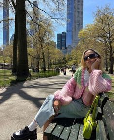 a woman sitting on top of a wooden bench in a park next to tall buildings