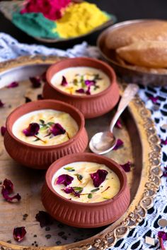 three bowls of soup on a plate with spoons next to bread and flower petals