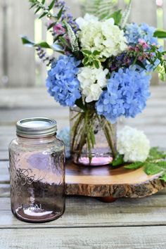 blue and white flowers are in a mason jar on a wooden tray next to a vase