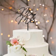 a white wedding cake with pink flowers and the word yes on top is surrounded by lights