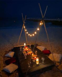 a table set up on the beach at night with candles and lights strung from it