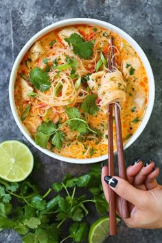 someone holding chopsticks over a bowl of noodles with cilantro and lime