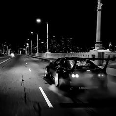 a black and white photo of a car driving down the road at night with city lights in the background