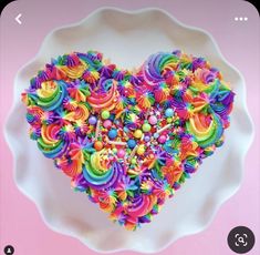 a white plate topped with a heart shaped cake covered in rainbow colored icing and sprinkles