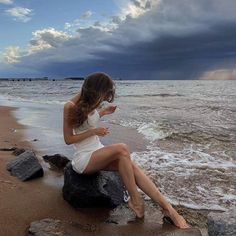 a woman sitting on top of a rock next to the ocean holding a cell phone