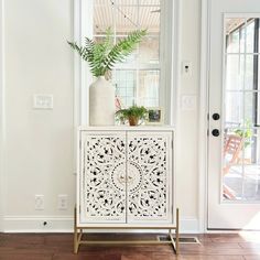 a white cabinet sitting in front of a window next to a plant on top of a wooden floor