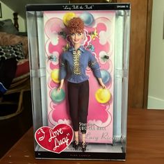 a barbie doll in a box on a table with a heart shaped sticker next to it