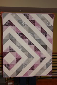 a person holding up a quilt made with squares and strips in pink, grey and white