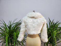 a white fur jacket sitting on top of a mannequin's head next to plants