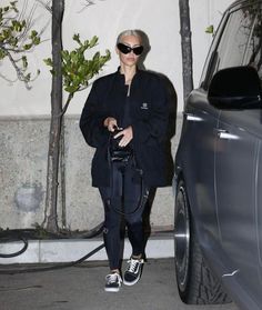 a woman in black jacket and leggings walking next to a car with sunglasses on