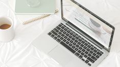 an open laptop computer sitting on top of a white bed next to a cup of coffee