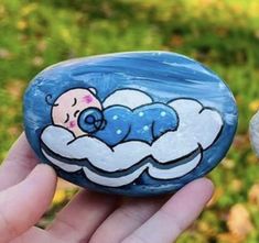 a hand holding a painted rock with a sleeping baby on it's back in the grass