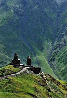 an old building on the side of a mountain with green grass and mountains in the background