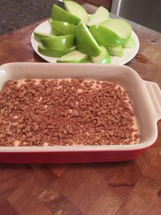 a red dish filled with food sitting on top of a wooden table next to green apples