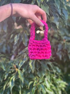 a hand holding a pink crocheted purse in front of a bush with green leaves