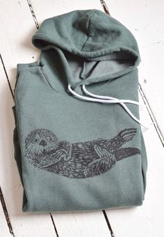 This super soft sponge fleece pullover hoodie is hand printed with our original Otter design in our home studio on the Coast of Maine. It features kangaroo pockets, ribbed cuffs and waistband, has a hood drawstring and is pre-shrunk. Details: * Original Hearth and Harrow Otter design, printed in black * Bella + Canvas Sponge Fleece Hoodie in green * Eco-friendly, water-based ink * Unisex sizing * Machine wash cold, tumble dry The handmade quality of our products assures that each one is unique. Cute Sea Otter, Dark Green Hoodie, Otter Cute, Otter Design, Hoodie Cute, Sea Otter, Aesthetic Shoes, Green Hoodie, Fashion T Shirt