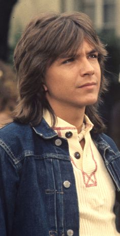 a man with long hair wearing a denim jacket and looking off to the side in front of a building