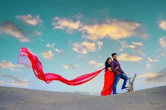 a man and woman standing on top of a sandy hill holding a red scarf over their heads