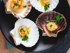 four scallop shells on a black plate with parsley garnishes