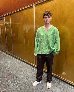 a young man standing in front of a gold wall wearing a green sweater and black pants