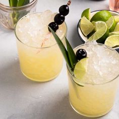 two glasses filled with lemonade, limes and olives on a counter top