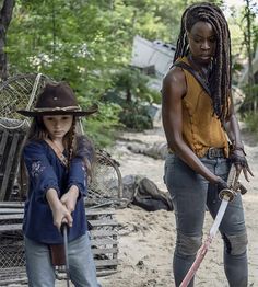 two women in cowboy hats holding swords on a dirt road with trees and buildings behind them