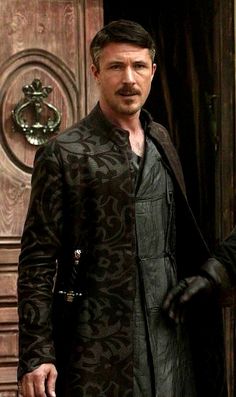 a man with a moustache standing in front of a door wearing a coat