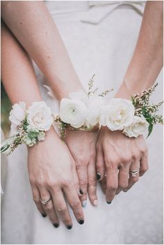 two brides holding hands with white flowers on their wristbands in front of the camera