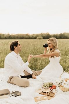 a man and woman sitting on a blanket in the middle of a field with food