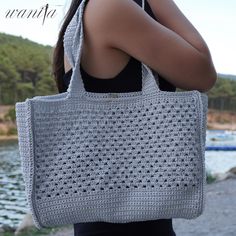 a woman is carrying a crocheted tote bag by the water with her hands behind her back