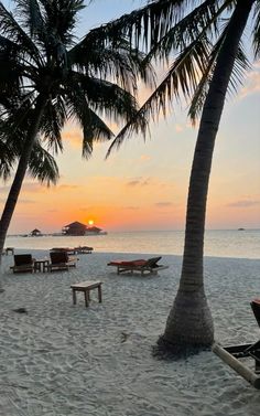 the sun is setting behind two palm trees on a beach with chairs and tables in the sand