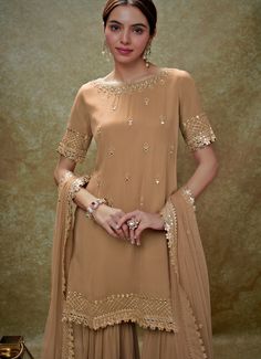 This exquisite Beige Georgette Kurta Sharara Set is a perfect blend of elegance and tradition. Made from georgette fabric, the beige kurta is adorned with delicate embroidery featuring mirrors, gota, sequins, and pearls, creating a stunning visual appeal. The neckline, hem, and sleeves are intricately embellished, complemented by small floral buttis that add a touch of grace. Teamed with a tiered sharara that showcases scattered mirror work, this ensemble exudes sophistication. The look is completed with a matching net dupatta, beautifully bordered and dotted with buttis, making it an ideal choice for Home Pooja, family gathering or festive occasions. Composition : Kurta and Sharara - Viscose Georgette and Dupatta - Soft net Care: Dry Clean Only and Vacuum Storage This product can be custo Mirror Work Kurta, Bridal Crop Top, Beige Kurta, Kaftan Kurta, Georgette Kurta, Kurta Lehenga, Prince Coat, Kurta Sharara Set, Waistcoat Woman