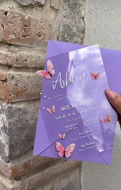 a person holding up a purple wedding card with butterflies on the front and back, in front of a brick wall