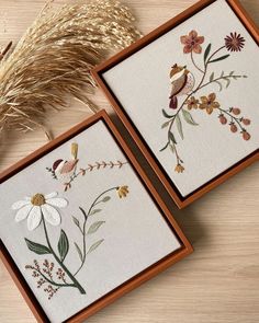 two framed pictures with flowers and birds in them on a wooden table next to some dry grass