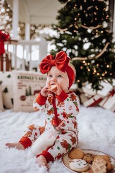 a baby sitting in front of a christmas tree eating cookies and looking at the camera