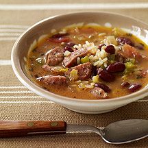 a bowl of soup with beans, sausage and rice on a tablecloth next to a spoon