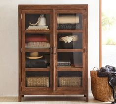 a wooden cabinet with glass doors and baskets on the bottom shelf, next to a wicker basket