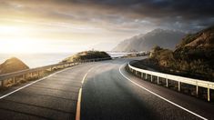 an empty road near the ocean with mountains in the backgrouds and sun shining down on it