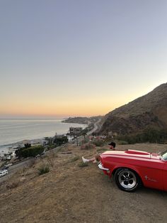 a red car parked on the side of a hill next to the ocean at sunset