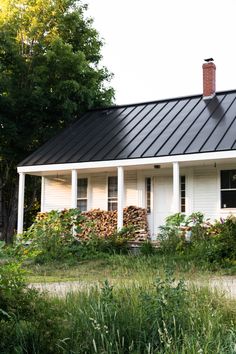 a small white house with a black metal roof and chimney in the front yard surrounded by tall grass