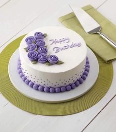 a white cake with purple flowers on it sitting on a plate next to a knife and fork