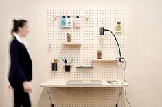 a woman walking past a desk with a laptop on it and shelving behind her