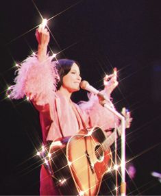 a woman holding a guitar and singing into a microphone while standing on stage with her arms in the air