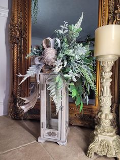 a white lantern with greenery in front of a mirror and lamp on the floor