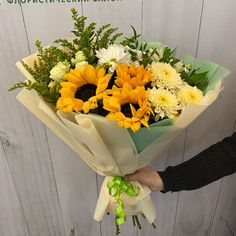 a person holding a bouquet of sunflowers and greenery in their hand, against a white wooden wall