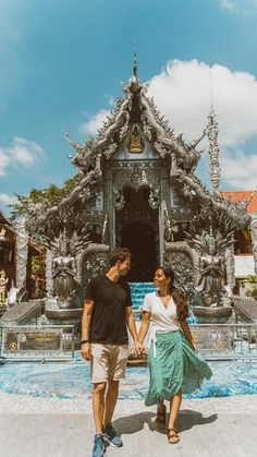 a man and woman holding hands in front of a fountain with statues on the sides